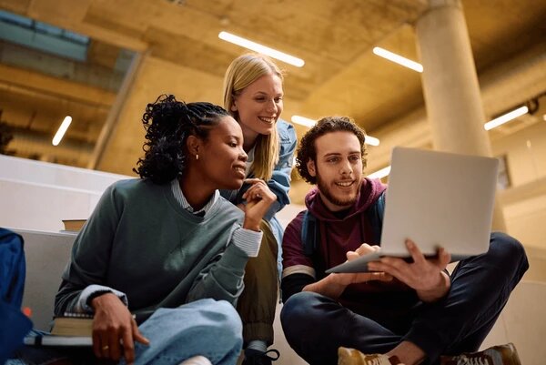 Three students smiling and looking at a laptop together while sitting on steps in a school building